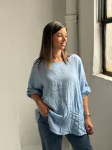 Blusa celeste de corte holgado, con cuello redondo, mangas tres cuartos con trabilla y detalles de alforzas en el frente.