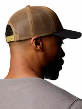 Gorro trucker Columbia Mesh Snap Back Hat, color marrón tostado con visera gris oscura y paneles traseros de malla. Cuenta con un parche gráfico redondo en el frente con diseño de montañas y cierre trasero ajustable.