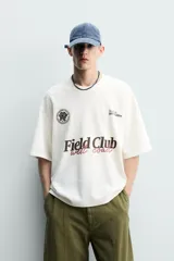 Remera de algodón color crudo, con textura de tejido abierto, cuello redondo y mangas cortas. Presenta estampas en contraste en el frente con los textos 'Field Club', 'West Coast', gráficos deportivos y un escudo circular.