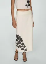 Falda midi de tiro alto con corte evasé, confeccionada en una mezcla de algodón y lino color beige. Presenta un diseño bordado floral en color negro ubicado en el lateral inferior.