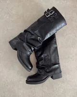 Bota alta de caña arrugada, color negro, confeccionada en cuero vegano. Presenta un taco bajo y hebillas decorativas en la parte superior de la caña.