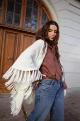 Poncho tejido de color blanco, con flecos en los bordes y detalles de bordado en crochet a lo largo de la abertura frontal. Cuenta con un cierre de cordones con borlas en el cuello.