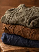 Buzo de lana merino color marrón con diseño de tejido trenzado en el frente y cuello redondo acanalado. Presenta puños y ruedo elásticos, con una pequeña etiqueta decorativa en la parte inferior.