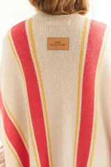Ruana artesanal de tejido de punto, color beige con franjas horizontales gruesas rojas y finas amarillas. Posee flecos en el borde inferior.