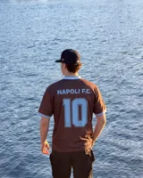 Camiseta de fútbol color marrón con cuello en V y detalles en celeste, inspirada en el Napoli F.C., con diseño de mangas cortas y sponsors impresos en el frente.