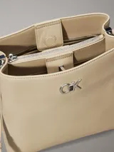 Cartera tipo saco de piel sintética estructurada color beige, con correa ajustable para el hombro y herrajes plateados. Presenta el monograma de Calvin Klein en la parte frontal.