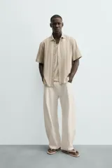 Camisa de calce relajado confeccionada en tejido con mezcla de algodón y lino, con diseño de rayas verticales en tonos beige. Presenta cuello bowling, manga corta, aberturas laterales en el bajo y cierre frontal con botones.