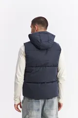 Chaleco puffer acolchado color azul marino, con capucha fija, cierre frontal con solapa de botones a presión y bolsillos laterales con cierre. Presenta un parche con el logo de la marca en el pecho.