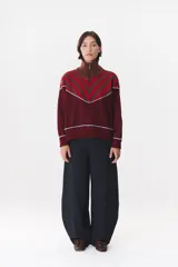 Buzo de tejido de punto en color azul marino, con cuello alto y cierre frontal parcial. Presenta un diseño con franjas en color marrón y detalles de líneas en rojo en el pecho, hombros y ruedo. Tiene corte holgado y mangas largas con puños acanalados.