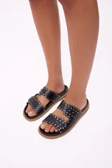 Sandalia tipo mule de mujer, color dorado metalizado, con dos tiras anchas superpuestas con borde festoneado y tachas doradas. La suela es gruesa y de color marrón claro.