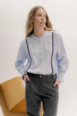 Camisa de algodón blanca con cuello a la base, cierre frontal con botones y mangas largas con puños delicados. Presenta volados verticales y detalles de cintas de terciopelo en contraste a lo largo del frente.