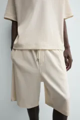 Bermuda de corte relajado, color crema o blanco roto, con textura acanalada (canalé). Presenta cintura elástica ajustable con cordón, bolsillos de vivo frontales y un bolsillo plastrón trasero.