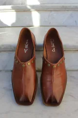 Zapato de cuero vacuno color bordó, con diseño de punta cuadrada y detalle de tachas metálicas planas en el borde superior. Presenta un taco geométrico bajo y costura central en el empeine.