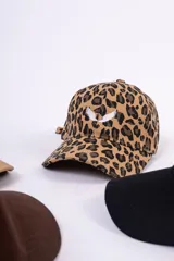 Gorra tipo baseball con estampado animal print de leopardo en tonos marrones y negros. Presenta un bordado frontal en color blanco con forma de alas y cierre posterior ajustable.