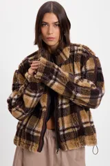 Campera de corderito sintético con diseño a cuadros en tonos marrones y negros. Presenta cuello alto, cierre frontal completo y detalles de paneles en color marrón liso. El diseño es de silueta holgada, largo tipo crop y cuenta con un cordón elástico ajustable en el ruedo.