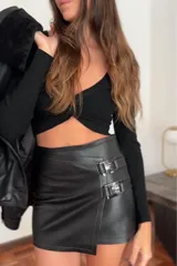 Minifalda negra de cuero sintético, tiro bajo, con corte irregular y detalle de dos hebillas plateadas en el lateral.