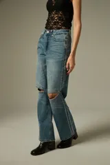 Pantalon de jean azul con corte recto y ancho, presenta aberturas en las rodillas y detalles de tachas metálicas a lo largo de las costuras laterales.