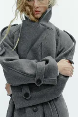Abrigo largo gris de corte cruzado con solapas anchas, doble botonadura y cinturón ajustable en el mismo tejido. Cuenta con mangas largas con trabillas en los hombros y puños, además de bolsillos delanteros con solapa.