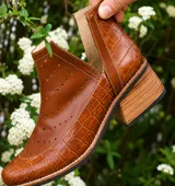 Botín de cuero blanco con textura de crocco y charol, diseño con calados circulares y aberturas laterales en forma de V. Cuenta con taco foliado de madera y vira cosida.