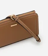 Cartera bandolera pequeña de formato rectangular, confeccionada en poliuretano color tostado con costuras decorativas en contraste. Cuenta con cierre superior de cremallera con tirador alargado, placa metálica frontal con logo y correa larga ajustable y desmontable mediante mosquetones.