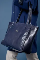 Bolso tipo shopper de gran tamaño confeccionado en cuero vacuno color marrón oscuro. Presenta laterales con textura grabada tipo cocodrilo y detalles de costuras decorativas con tientos de cuero en los paneles frontales y traseros. Incluye doble asa de hombro, cierre superior y placa metálica decorativa en el frente.