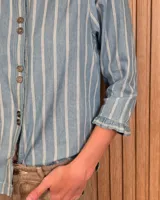 Camisa de algodón color blanco con rayas verticales rosadas, cuello alto con volados, mangas tres cuartos abullonadas con puños elastizados con volados y cierre frontal de botones oscuros.
