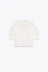 Blusa de algodón color blanco con cuello redondo y mangas abullonadas. Presenta detalles de puntilla a tono en el pecho y las mangas, bajo recto y cierre posterior con abertura y botón.