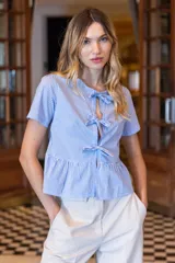 Camisa celeste a rayas verticales blancas, con cuello redondo y mangas cortas. Presenta abertura frontal con lazos decorativos y peplum fruncido en la cintura.