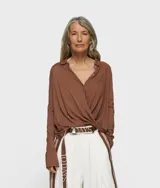 Blusa cruzada de punto de algodón orgánico color marrón, con cuello, cierre de botón, puños con abertura y monograma bordado en la manga izquierda. Tiene bajo recto y aberturas laterales.