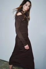 Vestido largo de color marrón oscuro, con cuello fluido tipo bote, manga larga y detalle de fruncido lateral con broche metálico en la cintura.