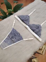 Conjunto de lencería compuesto por un corpiño triangular y una bombacha tipo tanga, confeccionados en encaje gris con detalles de terminaciones en color champagne.