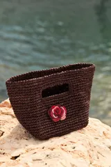 Mini bolso de mano tejido en rafia color marrón oscuro, con diseño estructurado y detalle de flor decorativa en tonos rojizos realizada en crochet.