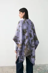 Ruana tejida de punto con diseño teñido artesanalmente en tonos lila y marrón, con terminación de flecos en el ruedo y etiqueta de cuero sintético en el frente.