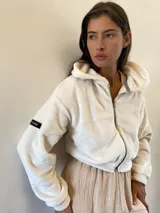 Campera corta de peluche en color blanco, con cierre frontal completo y capucha. Presenta un diseño de corte holgado y una etiqueta decorativa en la manga.