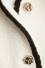 Chaqueta corta de tejido bouclé blanco, con ribetes negros en cuello, frente, bolsillos y puños. Presenta cuello en V, cierre frontal con botones dorados con diseño de nudo y bolsillos insertados.