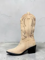 Botas estilo texano de gamuza en color beige, con diseño de costuras decorativas y tachas metálicas en la caña. Presentan punta fina y taco cuadrado en contraste de color negro.