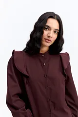Camisa de color bordó con cuello mao y cierre frontal de botones. Presenta volados en los hombros que aportan volumen y un diseño sofisticado.
