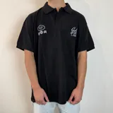 Chomba negra de corte oversize con cuello clásico y botones. Presenta estampados en el frente: un logo con las letras VCP y la palabra Golf, y un diseño con la frase "Golf Club".