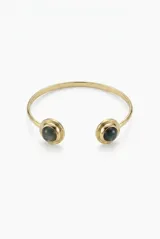 Pulsera rígida abierta de metal dorado con diseño minimalista, rematada en ambos extremos por piedras circulares de color oscuro.