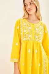Vestido largo hasta los tobillos de color amarillo, con bordados florales blancos a contratono. Presenta mangas tres cuartos, escote redondo con cartera de botones frontales y un diseño holgado.