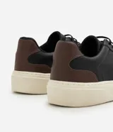 Championes casuales para hombre confeccionados en cuero negro, con diseño minimalista de caña baja, detalles en color marrón en el talón, cordones elásticos y suela alta de goma en tono off-white.