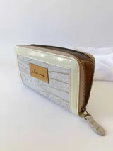 Billetera grande con cierre metálico, color beige y crema. El frente presenta un tejido texturado con patrón de rayas horizontales en tonos claros, ribetes en charol color crema y un parche de cuero marrón con la marca grabada.