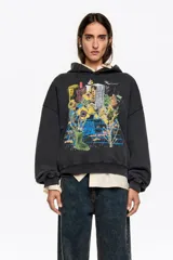 Sudadera con capucha de algodón color negro desgastado, corte globo y manga larga. Presenta un estampado frontal que muestra una escena urbana con edificios, flores amarillas y una bota de lluvia verde sobre un muro de ladrillos.