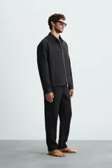 Sobrecamisa negra de corte regular, confeccionada en tejido de lino. Presenta cuello solapa, manga larga con puño abotonado y cierre frontal con cremallera metálica. Incluye bolsillos ocultos en las costuras laterales.