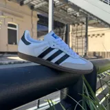 Championes Adidas modelo Samba ADV, de diseño clásico con base blanca, tres rayas laterales negras, puntera de gamuza gris y suela de goma color caramelo.