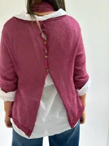 Sweater de punto color magenta con diseño asimétrico, cuello redondo y detalle de encaje bordado en el frente. Incluye una bufanda del mismo tejido y color, y se presenta superpuesto sobre una camisa blanca de algodón.