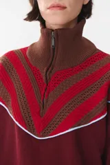 Buzo de tejido de punto en color azul marino, con cuello alto y cierre frontal parcial. Presenta un diseño con franjas en color marrón y detalles de líneas en rojo en el pecho, hombros y ruedo. Tiene corte holgado y mangas largas con puños acanalados.