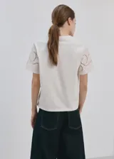 Camiseta color crudo, con cuello redondo y mangas cortas que presentan detalles bordados calados.