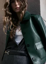 Chaqueta corta de efecto piel en color verde oscuro, con diseño recto, cuello camisero, manga larga y cierre frontal de botones ocultos. Presenta dos bolsillos de parche con solapa en la parte delantera y cuenta con forro interior.