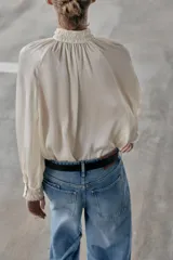 Blusa fluida de color blanco roto o crema, con cuello alto fruncido y manga larga con puños elásticos tipo nido de abeja. Presenta un frente abotonado con botones pequeños y brillantes.